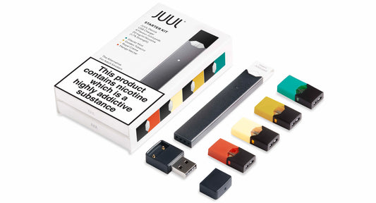 A JUUL starter kit showing a device, USB charger, and four pods in various colors. The box features a nicotine addiction warning, conveying caution.