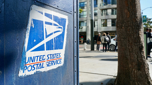Close-up of a USA blue postal service box with a white eagle logo on a city street. Background shows trees, pedestrians, and modern buildings.