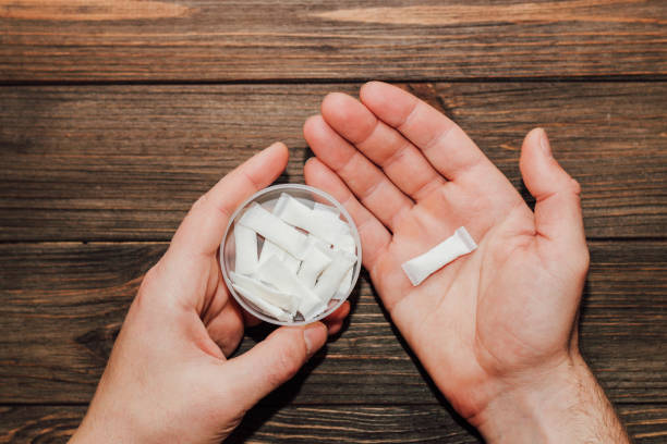 Two hands on a wooden table; one holds a clear bowl of white pouches, while the other holds a single pouch, suggesting tactile exploration of the items